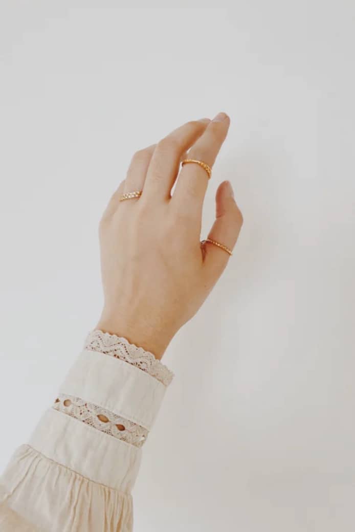 Close-up of a left hand with three gold rings, wearing a lace-sleeved blouse against a pale background.