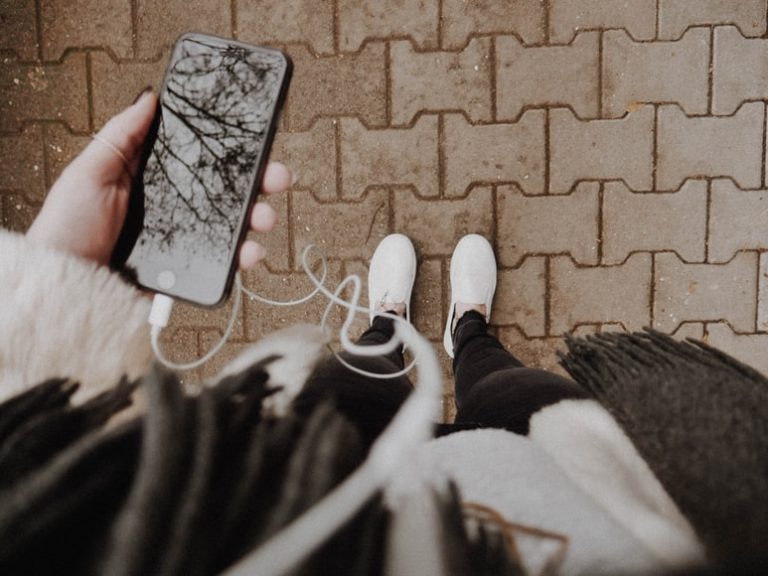 Top-down view of a person standing on a brick sidewalk, holding a smartphone with wired headphones plugged in.