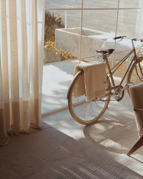 Bicycle indoors by large glass doors with a beige blanket draped over the saddle, sunlight streaming in from outside.